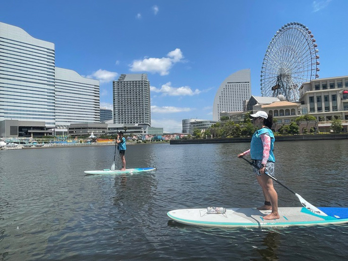 水辺荘のSUP体験ツアーに参加中の二人（みなとみらい地区を背景に）