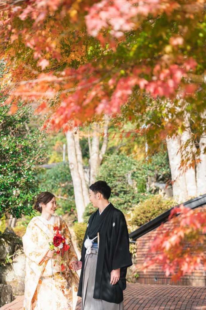 庭園の紅葉の下で微笑み合う和装の新郎新婦の写真